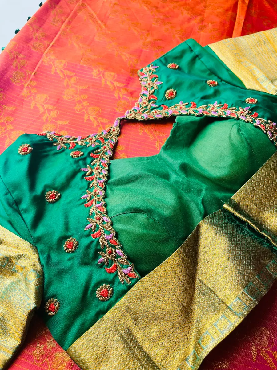 An emerald green blouse with pink and green aari embroidery worked along the neckline, laid against an orange silk.